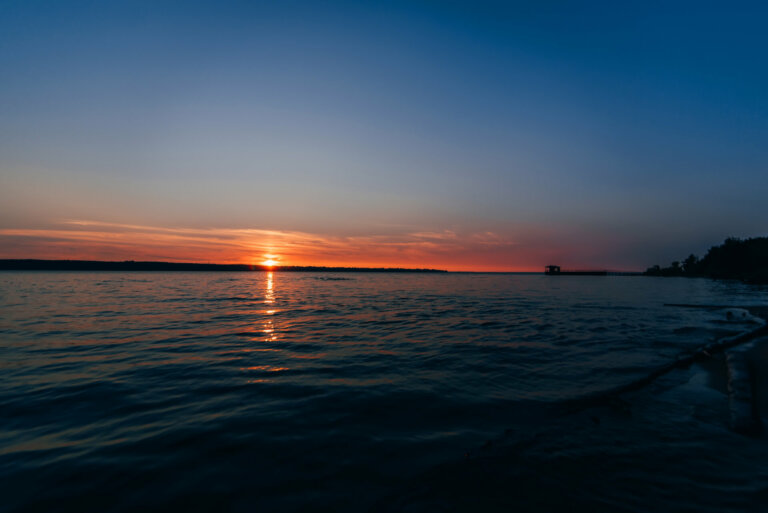 Un panorama côtier au coucher du soleil montrant l'intégration parfaite entre nature et technologie pour la résilience climatique. Des éoliennes offshore aux design bionique tournent doucement dans la lumière mauve du soir. Des barrières intelligentes de protection côtière émettent une lueur bleue apaisante. Des drones de surveillance maritime patrouillent, leurs capteurs créant des motifs lumineux sur l'eau. Au premier plan, une plage restaurée où des systèmes de surveillance de l'érosion s'illuminent doucement. L'horizon est ponctué de structures flottantes vertes pour la culture d'algues et la protection des écosystèmes marins.